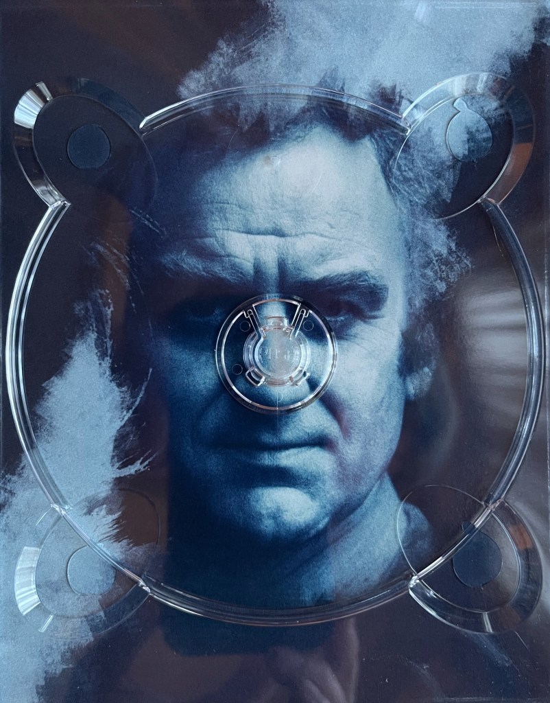 A blue-tinted image from one of the panels in the Blu-ray box set for The Sweeney Series 1, showing Regan facing straight on at us with a stern expression. It's behind a plastic holder for one of the Blu-ray discs, with the centre spindle lined up almost perfectly with his nose.