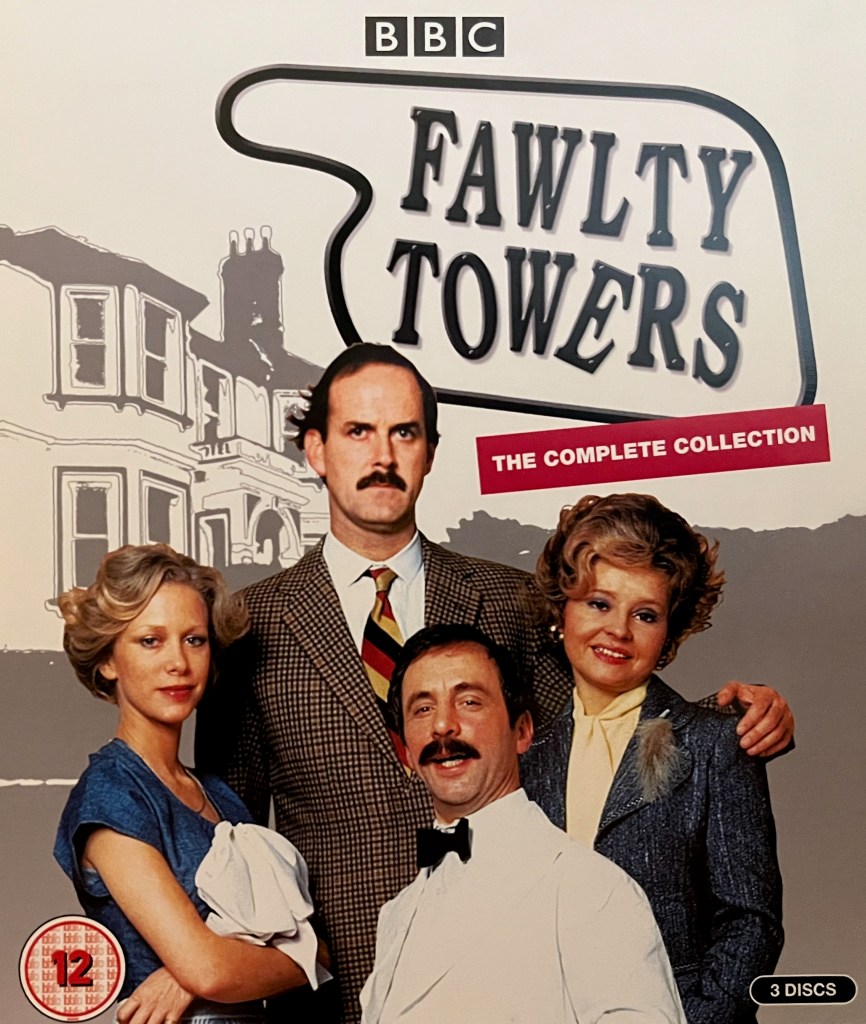 The Blu-ray cover art for Fawlty Towers, showing Basil, Sybil, Polly and Manuel standing together with the hotel in the background, a two-storey white building on a small hill. The white Fawlty Towers sign is shaped a bit like a pointing hand, aiming towards the hotel, with the black letters skewed as if they've individually been nudged around.