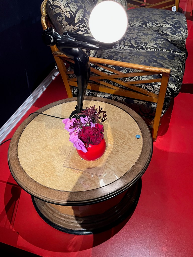 A black statuette of a woman leaning back with her arms outstretched together in front of her, forming the base for a lamp, the light of which is held in her outstretched hands. The lamp standing is on a round wooden table.