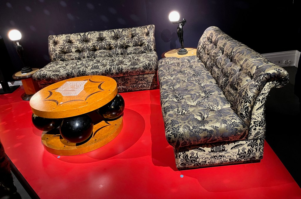 2 long sofas with a very leafy and ornate design on the upholstery. They are set at right angles to each other, facing a wooden circular coffee table, which instead of legs has 4 large black balls decorated with gold stars supporting it underneath, standing on a round wooden base.