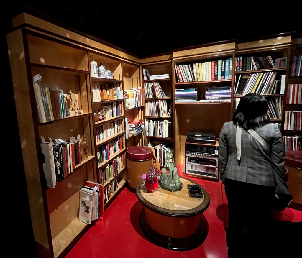 A huge bookcase with shelves in a variety of different sizes, filling the corner of the room. It is filled with books and records. In front of the bookcase is a round table with a compact CD player and an ornate table light with a sculpture of a person on it.
