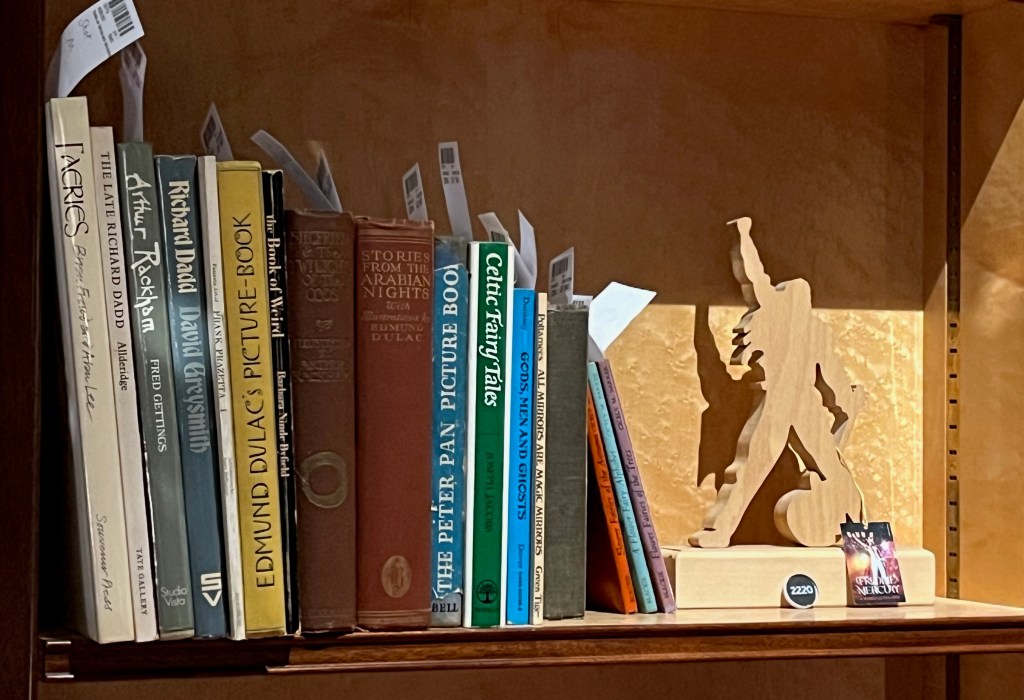 A collection of 17 fantasy and fairytale books, including titles about artist Richard Dadd, and books by illustrators like Edmund Dulac and C.M. Barker. Next to the books is a wooden 3D silhouette model of Freddie Mercury, standing with legs apart and one arm held in the air triumphantly.