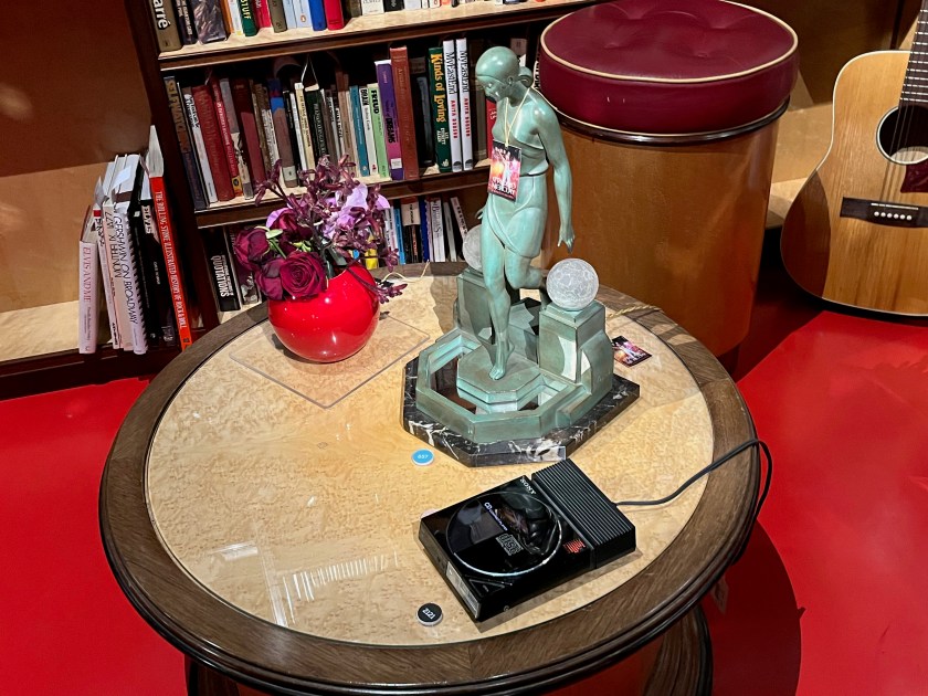 A round table holding 3 items. First is a round, red vase with colourful flowers. Next to that is an ornate table light that has a statuette of a woman stepping down from a platform between two plinths that are holding round lightbulbs. Then next to that is a Sony compact CD player.