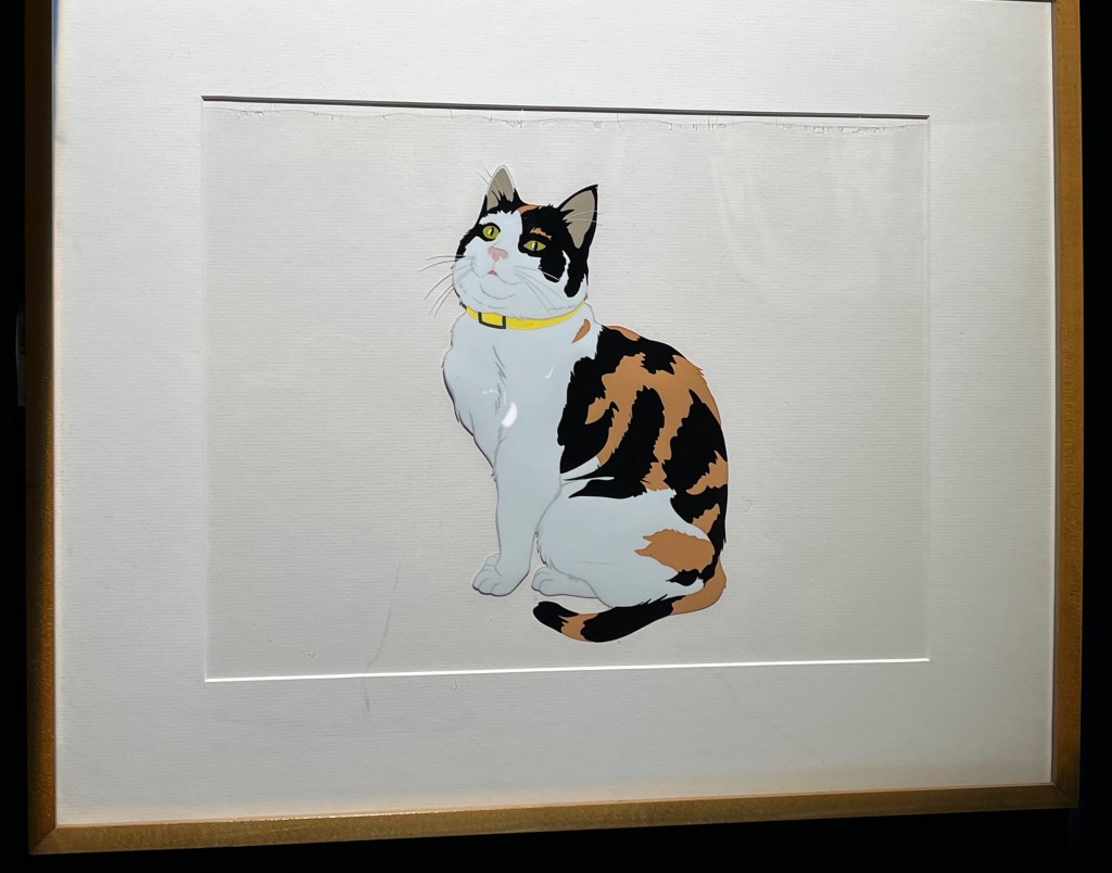 An animation cell by Walt Disney Studios of Freddie Mercury's cat Delilah, with brown and black striped fur on its back, tail and the top of its head, and white fur on its front. The cat is wearing a thin yellow collar and has a smiling face.