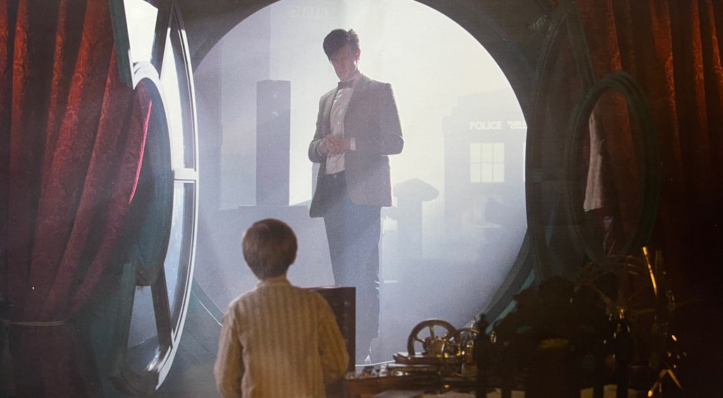 The reverse side of the cover sleeve for the Blu-ray of Doctor Who, A Christmas Carol. It shows Matt Smith's Doctor appearing in a large round doorway, facing the young boy Kazran Sardick, who has his back to us and is looking up at him 