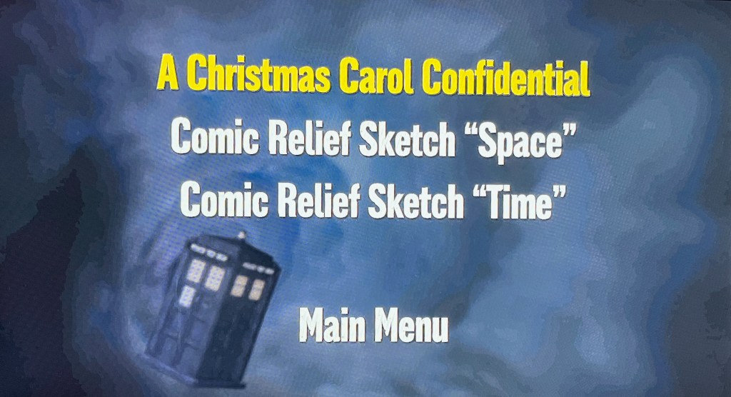 A screen from the audio navigation menu on the Doctor Who Series 6 Blu-ray. Against a static background of the swirling blue time vortex, with the Tardis in the bottom left, there is large, bold, white text forming the special features menu in the centre of the screen, with the options A Christmas Carol Confidential, Comic Relief Sketch Space and Comic Relief Sketch Time. The currently selected option is highlighted in yellow.
