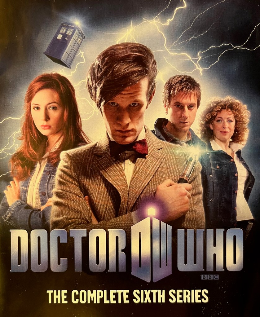 Front cover of the booklet from the old Blu-ray edition of Doctor Who Series 6, showing Matt Smith's Doctor holding his sonic screwdriver up against his left shoulder, while behind him are Amy, Rory and River Song. The Tardis is flying high above them in the distance. 