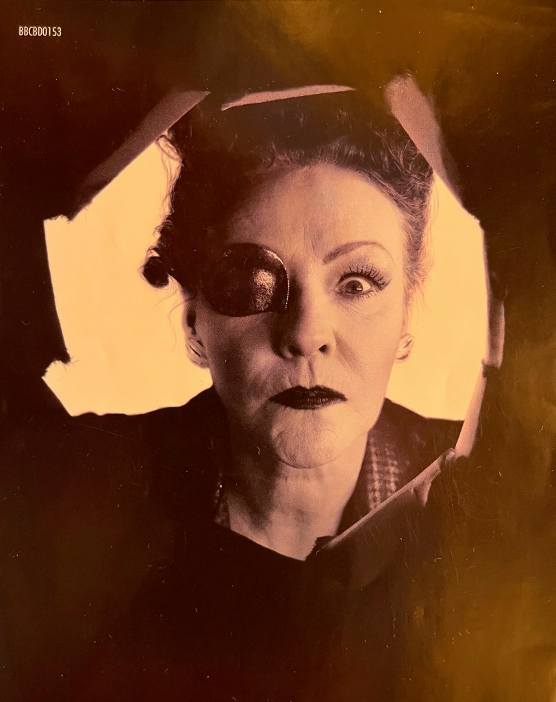 A page in the booklet from the old Blu-ray edition of Doctor Who Series 6, showing an image of Madame Kovarian, a stern looking lady with a patch over her right eye, looking through a large hole towards us.