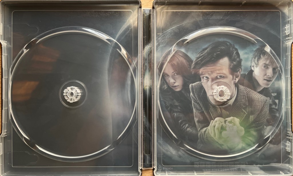 The interior of the Blu-ray steelbook for Doctor Who Series 6, when the discs have been removed. Behind the panels holding the discs is an image, on the right hand side, of Matt Smith's Doctor pointing his sonic screwdriver towards us, while Amy and Rory stand on either side of him.