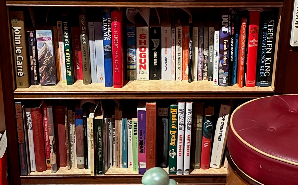 2 shelves in a bookcase, filled with novels on a comedy, science fiction and fantasy theme by authors like Tom Sharpe, John Le Carré and Stephen King, along with a variety of other books ranging from Freud on Sexuality to 2 copies of My East End by Anita Dobson.