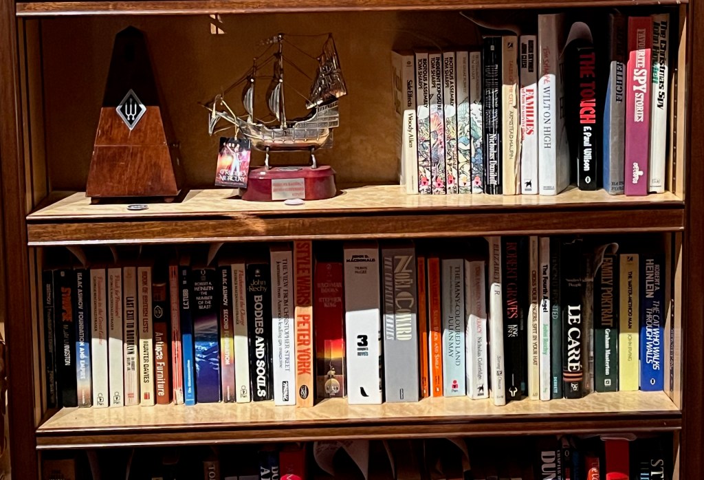 2 shelves in a bookcase, mostly filled with novels on a comedy, science fiction and fantasy theme by authors like Tom Sharpe, John Le Carré and Stephen King. Next to the books on the top shelf is a small model of the Santa Maria, a ship used by Christopher Columbus.