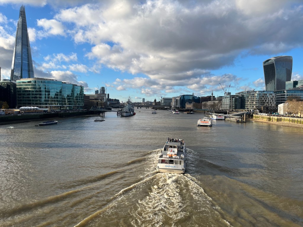 The view west along the River Thames from Tower Bridge as a boat travels into the distance. On the left is the South Bank, with the Shard looming tall over all the other buildings, while on the right is the north side of the river, with the so-called Walkie Talkie building looming large over those around it.