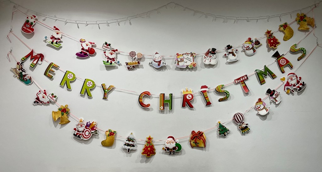 3 rows of Christmas bunting strung up on a wall. The top and bottom rows consist of various Christmas-related imagery, including Santa Claus and snowmen in various poses, stockings, bells and trees. The middle row consists of colourfully designed capital letters spelling Merry Christmas.