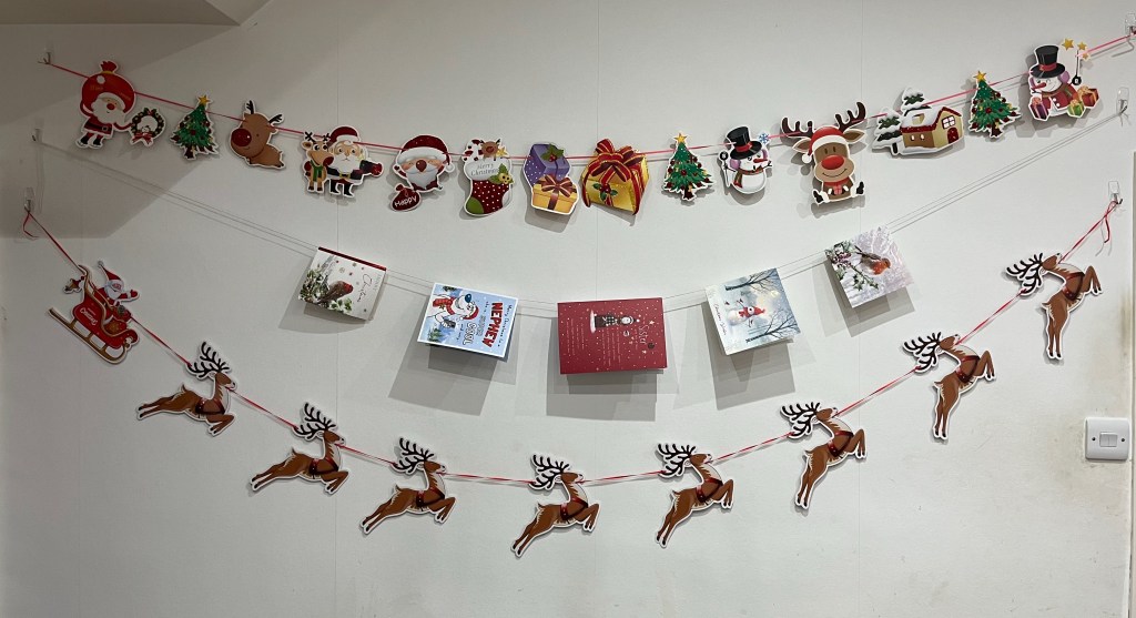 2 rows of bunting strung up on the wall. The top row has a variety of Christmas imagery including Santa, snowmen, presents, trees, reindeer and a snow-covered house. The bottom row consists of Santa in his sleigh being pulled along by 8 reindeer, which are spread along the ribbon so it looks like they're flying down towards the centre and then swooping up as they move across the wall. In between the two rows of bunting is a line of string on which 5 Christmas cards are hung on display.