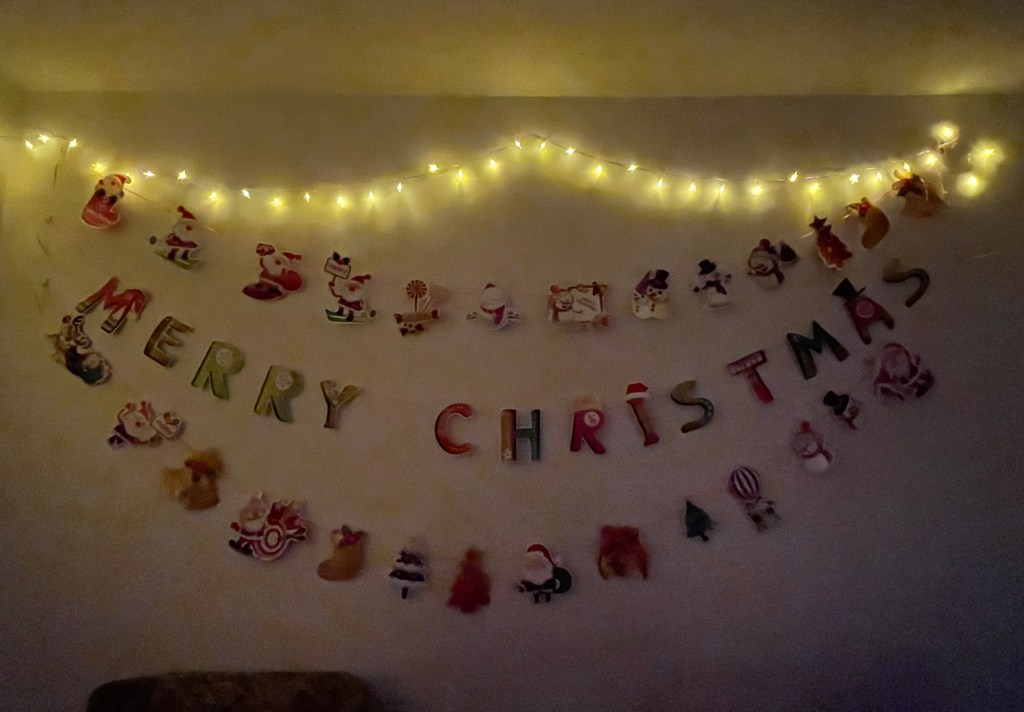 A string of fairy lights high on a wall above some Christmas bunting, which has lots of festive images and the text Merry Christmas in large letters.