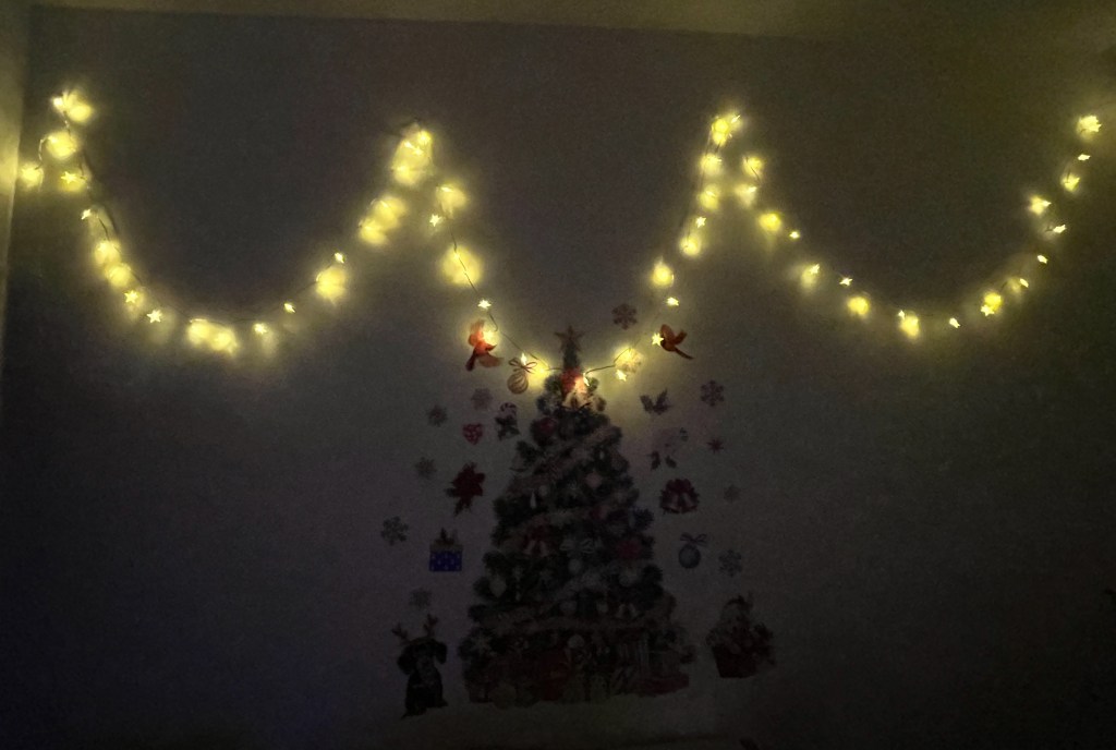 A string of fairy lights on a wall, curving down and up a few times, above some festive wall stickers including a large Christmas tree and lots of small festive images surrounding it.