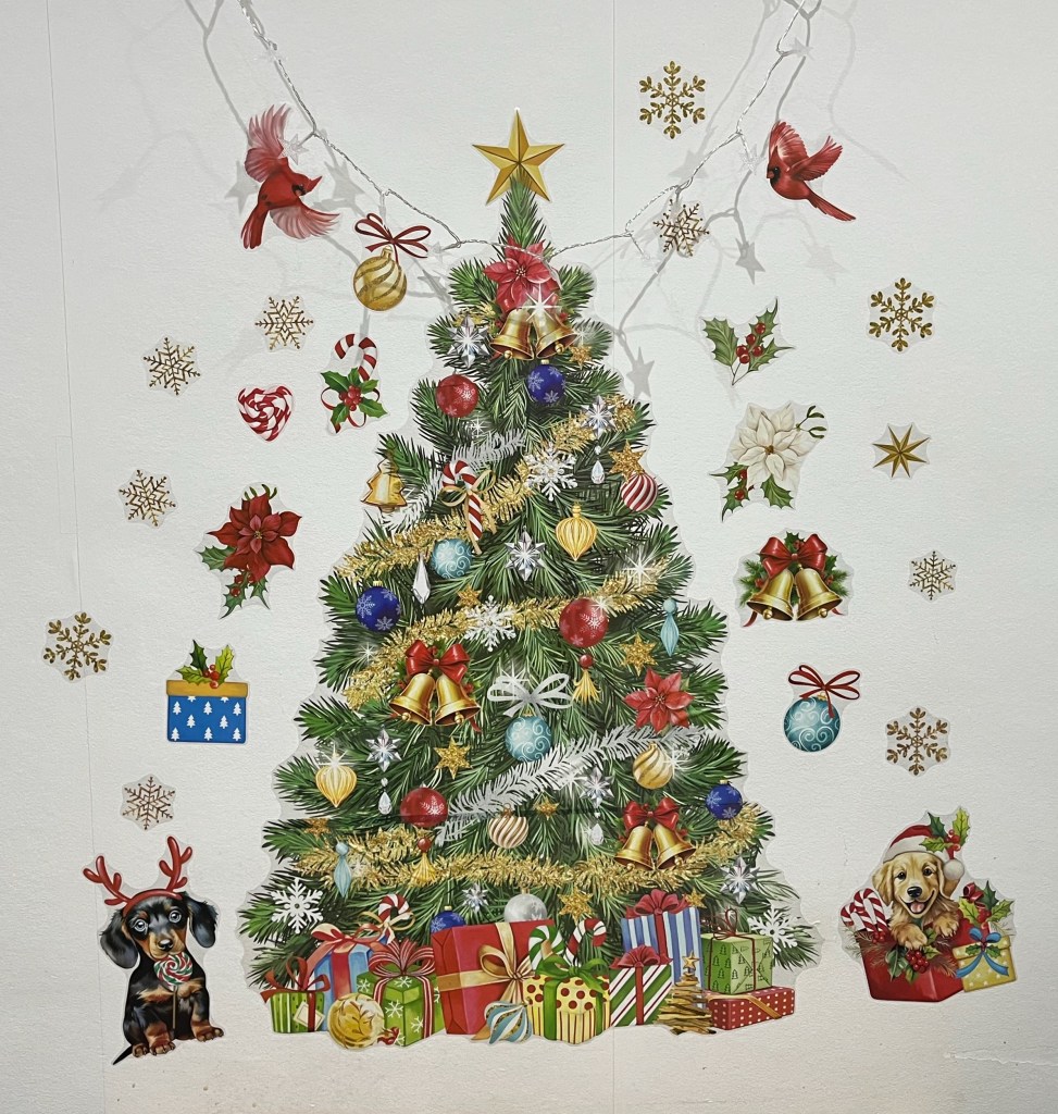 Christmas themed stickers on a plain white wall. In the centre is a huge Christmas tree, made up of 4 large stickers lined up one above the other. The tree is heavily and colourfully decorated with baubles, bells, snowflakes and striped candy canes. There is a star on the top, and a large array of presents in colourful wrapping at the base. Around the edge of the tree are lots of smaller sticks, including snowflakes, holly, bells, birds and baubles. Either side of the base of the tree are a couple of dogs, one with antlers on its head as it nibbles on a round lollipop, the other wearing a Santa hat and looking very happy as it sits up to look out of a red box with candy canes and a wrapped gift next to it.