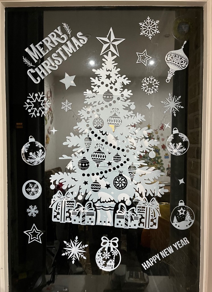 A window covered in a festive selection of white stickers. At the centre is a large tree made up of a few stickers lined up with each other. It has lights and decorations on it, and presents across the base. Around it are lots of smaller stickers, mainly of varying designs of snowflakes, baubles and stars, along with the text Merry Christmas at the top left of the window and Happy New Year in the bottom right.