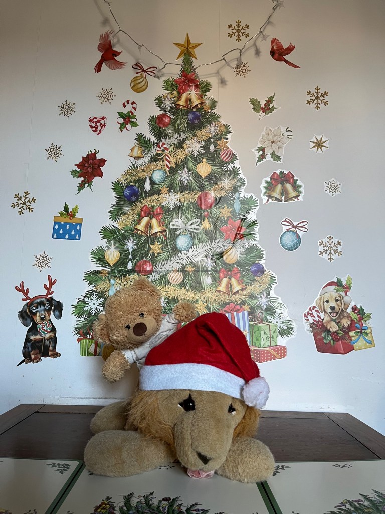 A large cuddly toy lion, wearing a red and white Santa hat and sticking its tongue out, is sat on a dining table. Sat on its back, looking over its shoulder, is a smiling teddy bear wearing a white t-shirt with the face of Freddie Mercury on it. Behind them on the wall are a large variety of colourful festive wall stickers. In the centre is a huge Christmas tree bedecked fully in colourful decorations including bells and baubles, while around the tree are snowflakes, bells, baubles, holly, birds and dogs.