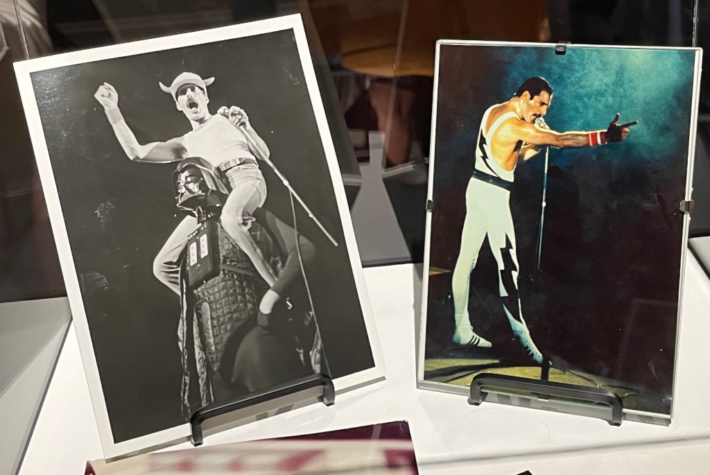 2 photos of Freddie Mercury. One, in black and white, shows him wearing a hat with horns as he rides on the shoulders of someone dressed as Darth Vader. The other, in colour, shows him singing on stage and as he points towards the crowd ahead of him.
