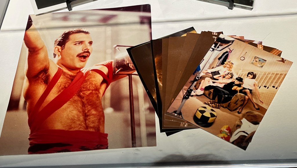 2 colour photos. One shows Freddie Mercury wearing a red strappy top as he sings and has his arms outstretched in the Radio Ga Ga video. The other shows the 4 members of a band crossdressed as women in a living room set for the video accompanying the song I Want To Break Free.