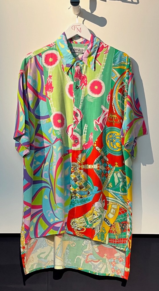 A brightly coloured short-sleeved shirt incorporating two different patterns, one in green, red, yellow and turquoise incorporating crowns, horses and saddlery motifs, the other in shades of fuchsia pink, lime green, purple and turquoise incorporating rearing horses and abstract patterns, cut slightly shorter at the front than the back, with white metal button fastening.