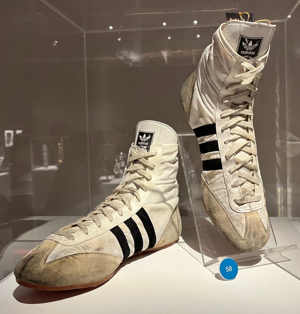 White cotton sneakers with black felt stripes and cream suede toes and heels. The Adidas brand logo is printed in black on the tongue of each shoe, and they have white laces.