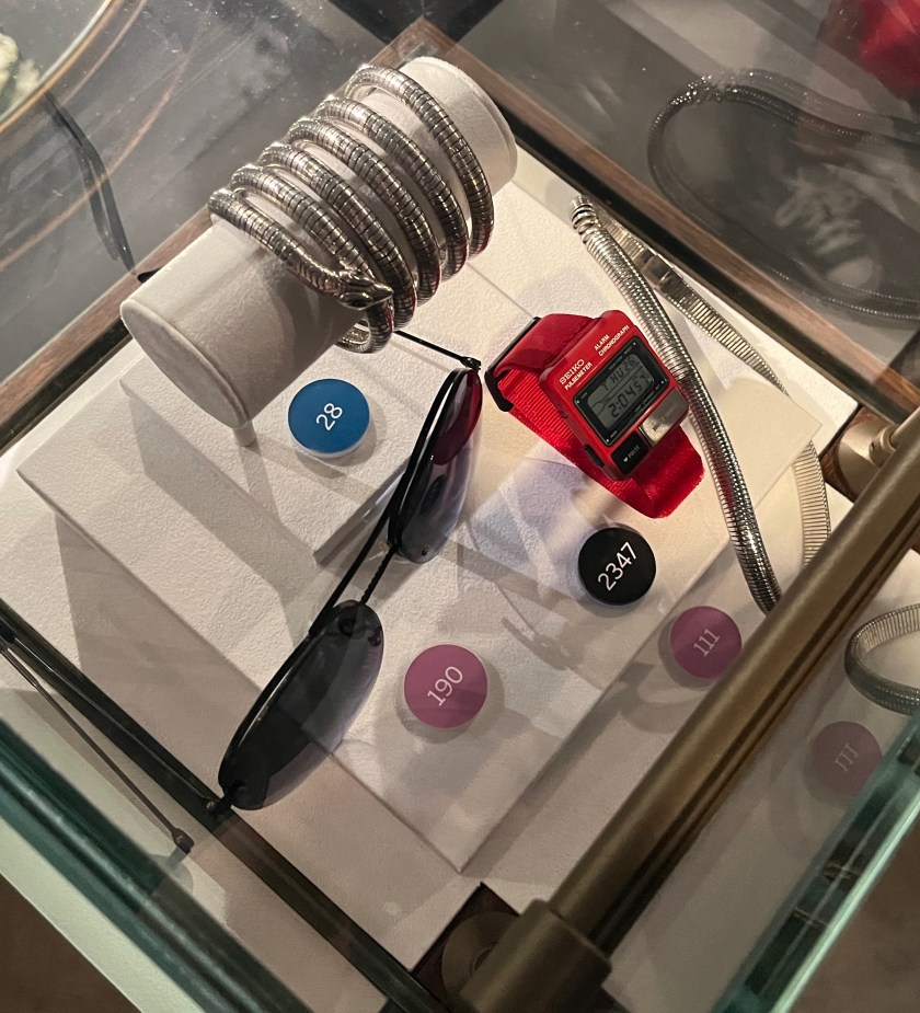 Top down view of a glass case containing a silver bangle designed like a snake, a silver choker, a pair of aviator sunglasses, and a red digital watch.