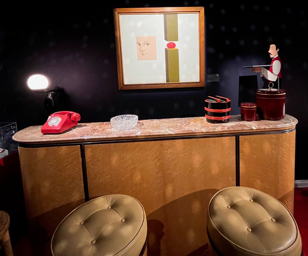 A drinks bar made of maple wood panelling with a pink marble top. Two bar stools sit in front of it, while on top of the bar is a telephone, ashtray and a few storage tins of varying sizes. A cardboard cutout of a waiter holding a tray stands behind the bar at the right end. A painting on the wall behind the bar shows a man's face looking towards an apple sitting on a wooden post, with another wooden post pointing downwards close above it.