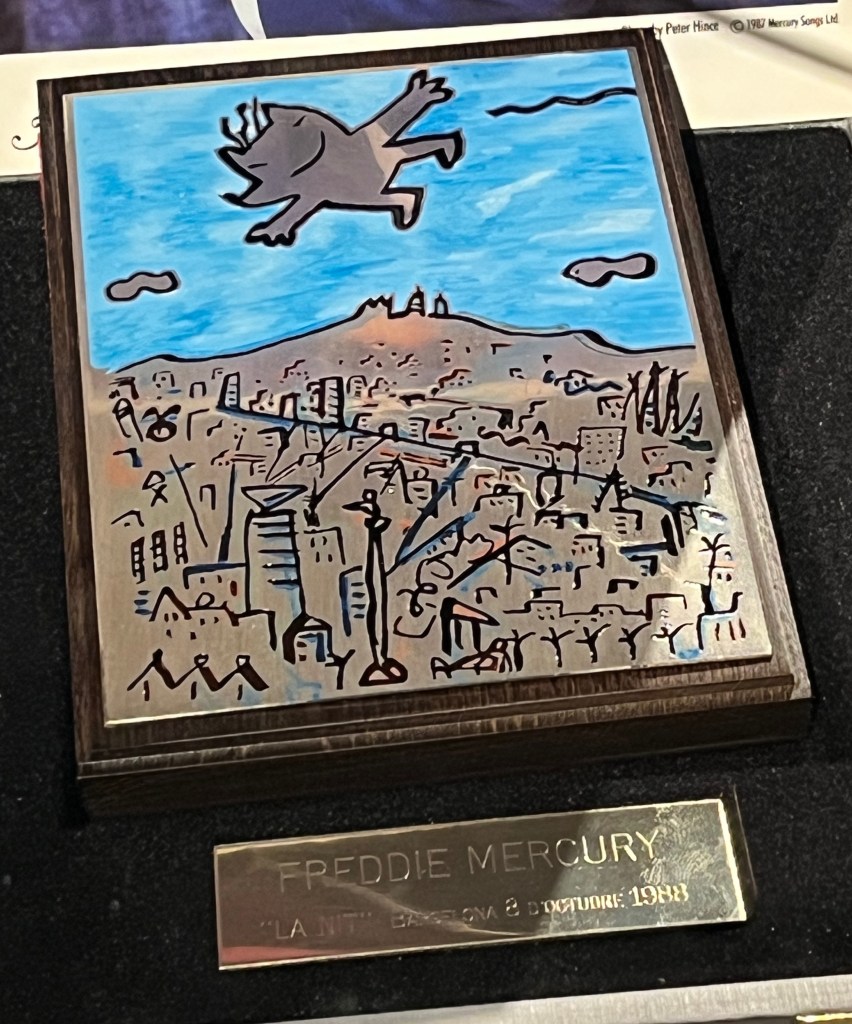A medal presentation box, with artwork of a grey creature with a smiling face, 2 arms and 2 legs flying over a mountainous cityscape. A silver plaque below the image is engraved with the text Freddie Mercury, La Nit, Barcelona, 8 October 1988.