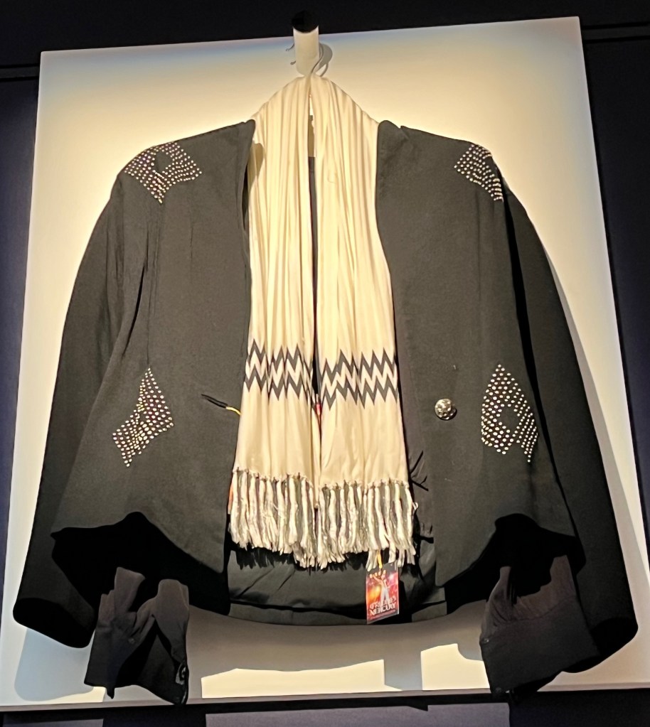 A white scarf with a double row of black zig-zag patterning at either end and black and white fringing. It's draped over a fine wool black jacket, the front decorated with four diamond shapes picked out in white metal studs. The collarless jacket also has padded shoulders, rounded lapels, fitted waist, raglan sleeves and single button fastening.