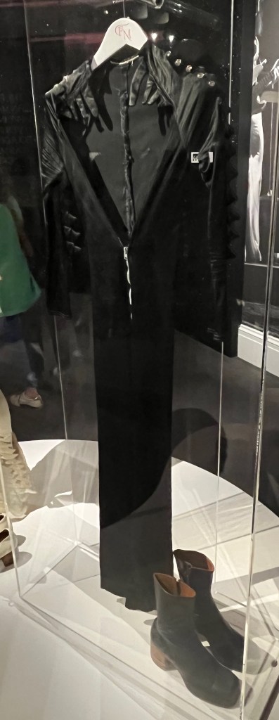 A catsuit of black stretch satin slashed to the waist at the front with angular, shaped neckline decorated either side at the top with several frond-shaped tabs. Both the deep cut front and long tight sleeves have zip fastenings. The suit also has slightly flared legs.