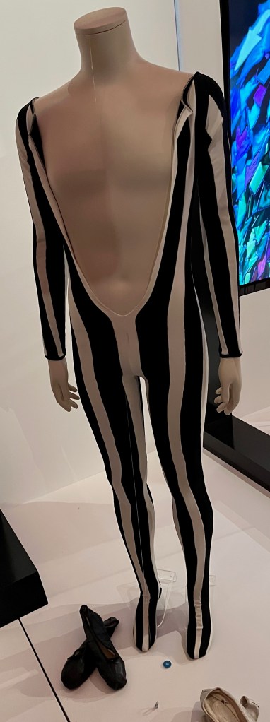 A black and white striped long sleeved elastane catsuit slashed to the waist, with integral feet. Each vertical stripe is a separate narrow panel stitched alongside its opposing colour, while the cuffs are edged in black and the soles of the feet are shaped entirely from black.