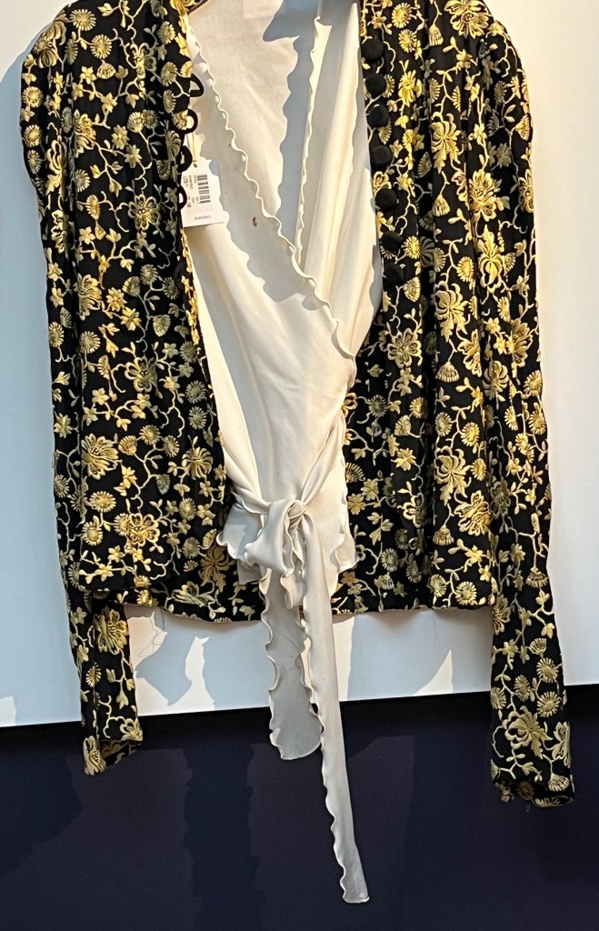 A black fitted jacket, pleated at the shoulders.The black shirt is heavily embroidered in yellow thread with a flower design. It fastens at the front with seven black fabric buttons and corresponding fabric loops. It's paired with a white nylon short-sleeve wrap-around top with a lightly frilled edge.