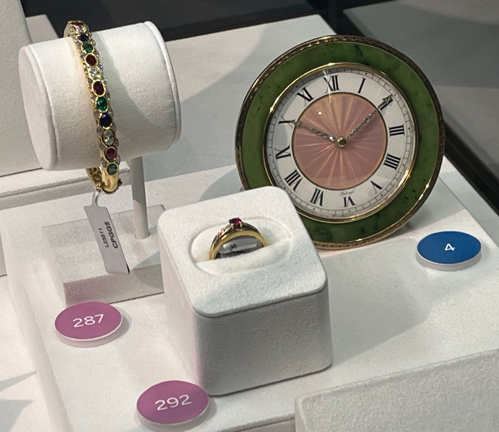 A ruby, emerald, sapphire and diamond bracelet, along with a ruby and diamond ring. Next to these is a Fabergé clock, where the numbers on the dial are black Roman numerals on a white background, but the centre of the dial is a translucent pink colour. There is a gold frame around the dial, and a wider green frame around the whole clock.