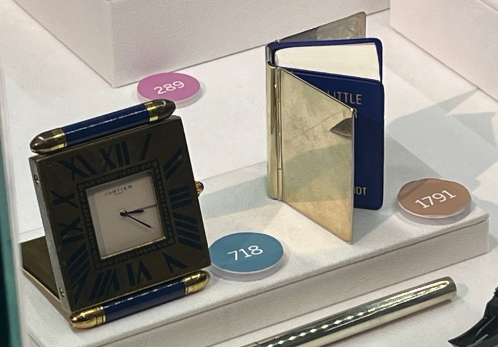 A small, square Cartier timepiece, with a white square dial inside a dark-coloured frame with black Roman numerals on it. Next to this is a tiny dictionary by Tiffany and co with a reflective silver cover surrounding an inner blue cover enclosing white pages.
