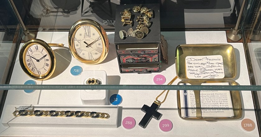 A glass case containing two Cartier timepieces, each consisting of a white face in a gold frame, along with an onyx and diamond ring, an onyx bracelet, a set of gold cufflinks, a black resin cross pendant, and a silver cigar case containing a hand written note and typed spiritual advice cards.