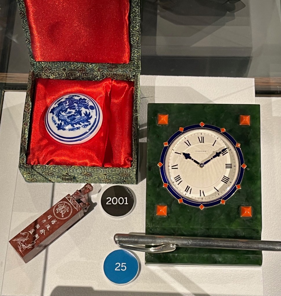 A small white stone desk seal, decorated in a blue Japanese design. This is next to a Cartier clock, consisting of a white clock face surrounded by a blue enamel border with small red diamond shapes marking each hour, all set into a rectangular green box with raised red squares near each corner, and a small cutout at the bottom for a pen to be laid in.