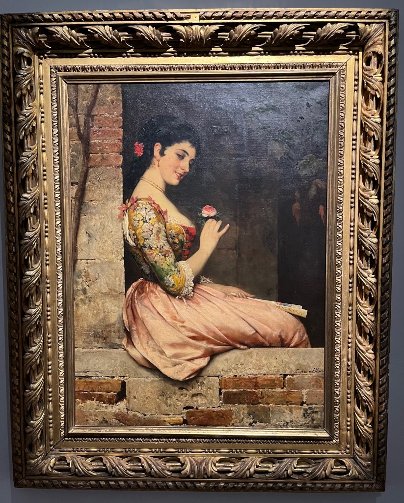 Oil on canvas painting by Eugen von Blaas called A Rose. It depicts a woman in a dress that has a colourful flowery top and plain skirt. She is sitting on a brick wall, leaning back against a taller brick wall behind her, smiling as she looks at a pink  rose she's holding in her hand.
