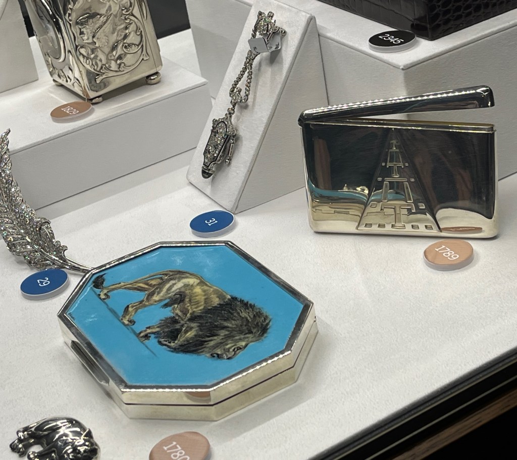 A ruby, sapphire, emerald, onyx and diamond fob watch, a diamond brooch in the shape of a feather, a silver card case with a triangle design, and an octagonal-shaped silver and enamel box with an image of a lion against a blue background.