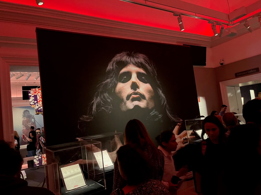 A gallery room in Sotheby's. Visitors are looking at pieces of paper in glass cabinets, overseen by a huge head and shoulders photo of Freddie Mercury, who appears to be glancing downwards as if watching over them. He is against a black background, his arms folded across his shoulders.