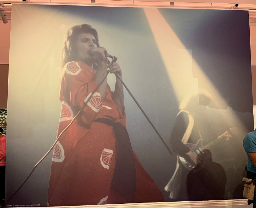 Freddie Mercury singing on stage while wearing a red kimono with a white pattern of stylised fans.