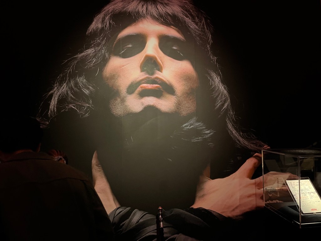 A head and shoulders photo of Freddie Mercury against a black background, his arms folded across his shoulders.