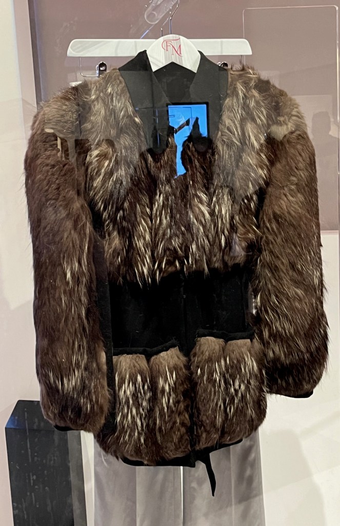 Half-length jacket with long pointed collar. The front is covered with fur from shoulders to chest, the pocket fronts and the upper half of the long sleeves, while there is also a black jersey tie belt that isn't furry.
