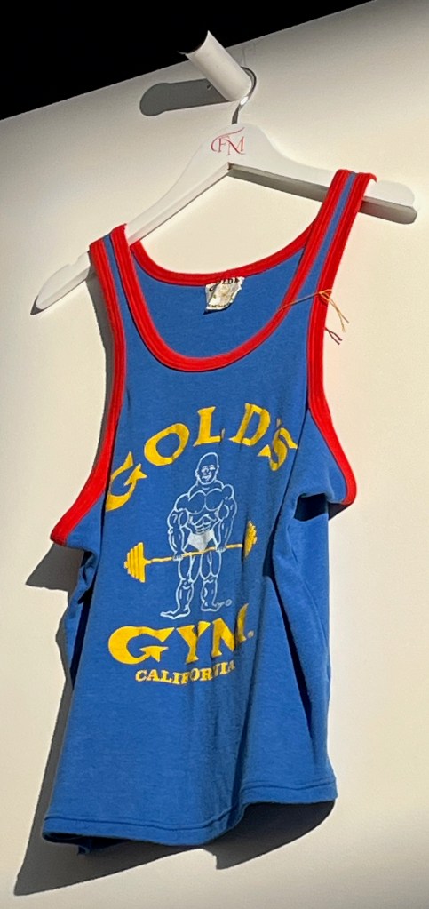 A blue cotton vest with red trim printed with yellow lettering ‘Gold’s Gym California’ and in white with a caricature of a weightlifter.