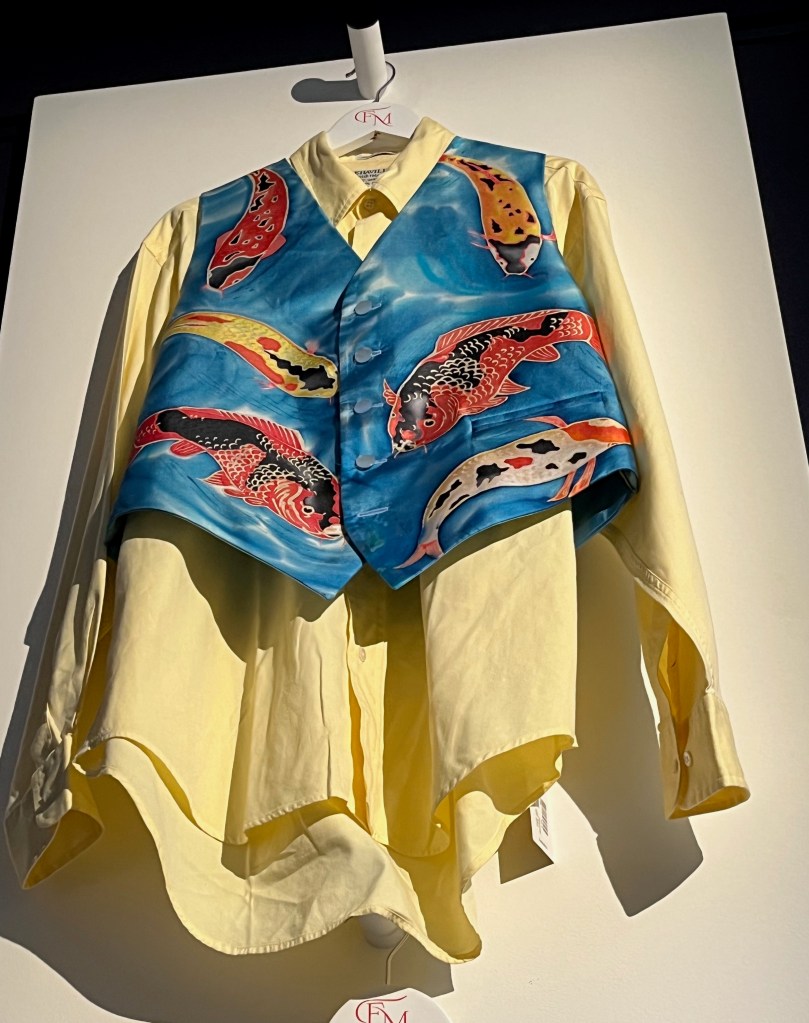 A hand-painted silk waistcoat with a design of Koi carp swimming in turquoise water, the carp in shades of orange, red, yellow and black with gold accents. It is on display over a yellow shirt.