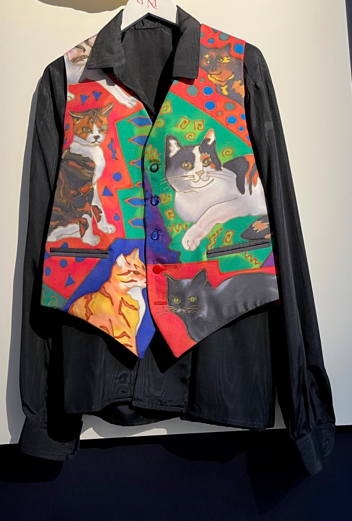 A silk waistcoat painted with a colourful design incorporating individual portraits of Freddie Mercury’s six beloved cats. It is on display over a black shirt.