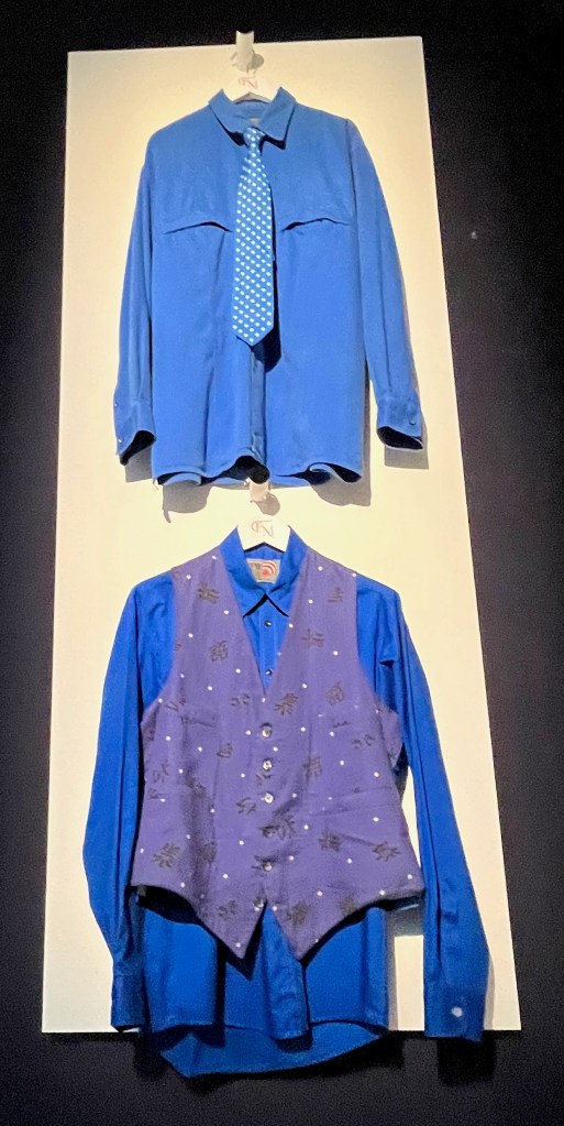 2 blue outfits hanging on a wall. At the top is an electric blue cotton shirt, with two breast pockets. This is paired with a blue and white polka dot silk tie. Meanwhile, on the bottom is a blue cotton waistcoat printed with white polka dots and black Japanese characters, with four slit pockets to the front. This is placed over a sky-blue cotton shirt with black buttons.