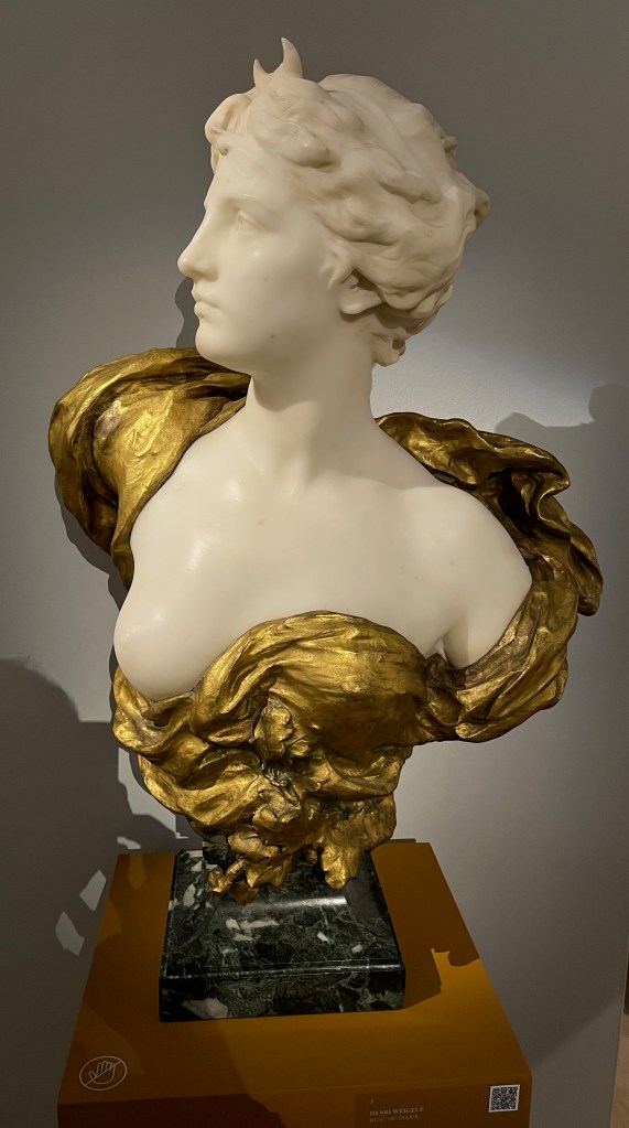 A white marble bust of the ancient Roman goddess Diana, showing her head and upper chest surrounded by an ornate gold design.