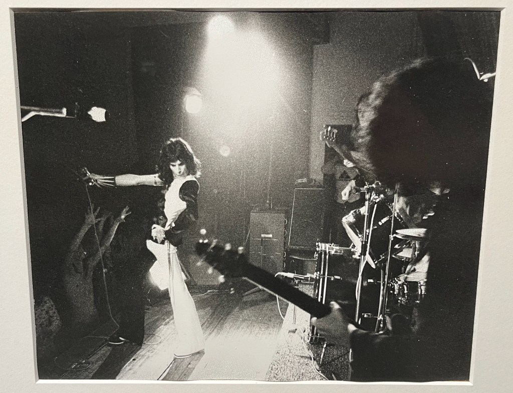 A black and white photo of Queen performing on stage at Imperial College in 1973, looking over Brian May's shoulder as he plays the guitar, towards Freddie posing with the microphone.
